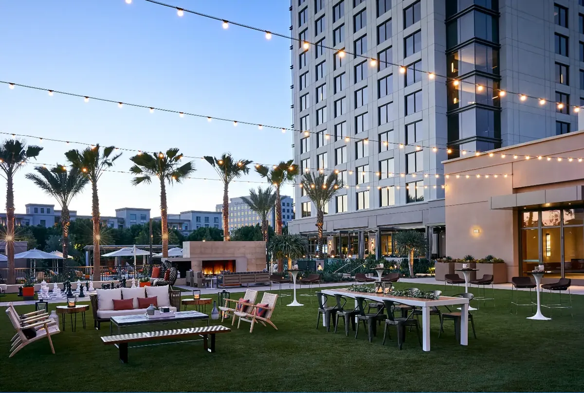 The outdoor lawn at the Marriott Irvine Spectrum at dusk, with string lights, palm trees, a lit fireplace, lounge seating and long communal dinner tables